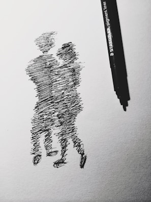A black ink drawing on white paper depicts two abstract human figures embracing or dancing. The sketching style is rough and dynamic, capturing a sense of movement and connection. A black pen is placed beside the drawing, suggesting the tool used to create it.