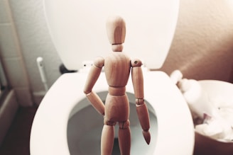 A wooden mannequin stands facing an open toilet seat in a bathroom setting. The scene is composed with the mannequin at the center, with the white toilet partially visible and other bathroom items slightly blurred in the background.