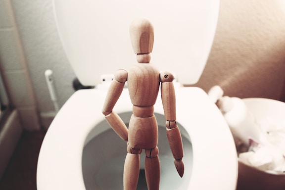 A wooden mannequin stands facing an open toilet seat in a bathroom setting. The scene is composed with the mannequin at the center, with the white toilet partially visible and other bathroom items slightly blurred in the background.