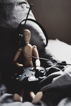 A wooden mannequin is sitting on a couch, holding a gaming controller. It appears to be wearing a headset, suggesting it's engaged in gaming. The background is composed of dark, soft fabric, creating a cozy and relaxed atmosphere.
