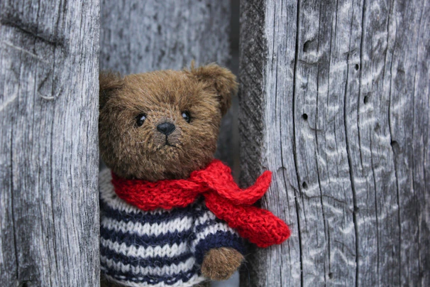 A cozy, smiling teddy bear wearing a tiny knitted scarf, sitting on a wooden shelf.