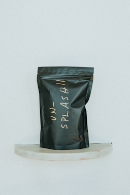 A black, matte-finished stand-up pouch with handwritten lettering in a light, cursive style is displayed against a minimalist background. The pouch is positioned on a small, semi-circular marble shelf and bears the word 'UNSPLASH!!' in an off-white color.