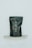 A black, matte-finished stand-up pouch with handwritten lettering in a light, cursive style is displayed against a minimalist background. The pouch is positioned on a small, semi-circular marble shelf and bears the word 'UNSPLASH!!' in an off-white color.