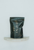 A black, matte-finished stand-up pouch with handwritten lettering in a light, cursive style is displayed against a minimalist background. The pouch is positioned on a small, semi-circular marble shelf and bears the word 'UNSPLASH!!' in an off-white color.