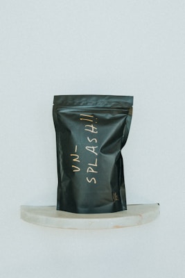 A black, matte-finished stand-up pouch with handwritten lettering in a light, cursive style is displayed against a minimalist background. The pouch is positioned on a small, semi-circular marble shelf and bears the word 'UNSPLASH!!' in an off-white color.