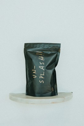 A black, matte-finished stand-up pouch with handwritten lettering in a light, cursive style is displayed against a minimalist background. The pouch is positioned on a small, semi-circular marble shelf and bears the word 'UNSPLASH!!' in an off-white color.
