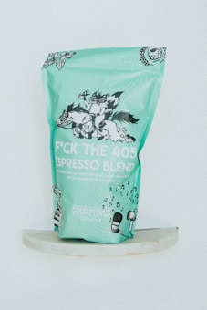A teal colored coffee bag stands upright, featuring whimsical black and white illustrations of animals in playful poses. The text on the packaging reads 'F*CK THE 405 ESPRESSO BLEND', accompanied by musical notes and decorative botanical elements.