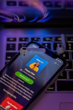 A smartphone displays a music streaming app screen titled 'Happy Hits!' along with a playlist description. The phone is placed on a laptop keyboard and two wireless earbuds rest on top. The lighting has a purple and blue hue, giving a modern and digital feel.