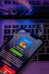 A smartphone displays a music streaming app screen titled 'Happy Hits!' along with a playlist description. The phone is placed on a laptop keyboard and two wireless earbuds rest on top. The lighting has a purple and blue hue, giving a modern and digital feel.