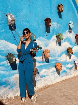 A person wearing a blue jumpsuit and sunglasses is smiling and engaged in a playful interaction with a series of vintage telephones mounted on a vibrant blue wall decorated with white cloud-like patterns. The wall features several multicolored telephones, each with a cord hanging down.