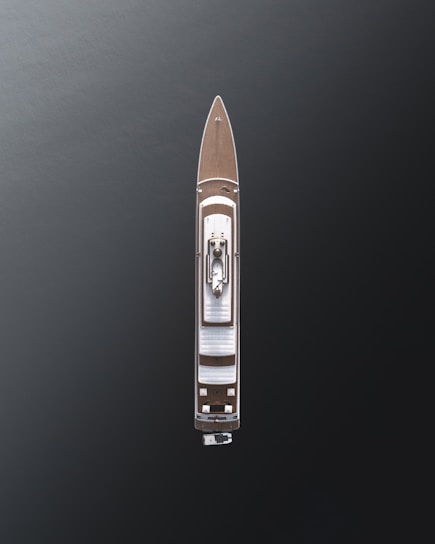 Aerial view of a large luxury yacht with a sleek design, positioned on dark, calm waters. The vessel features a wooden deck and a central structure, showcasing modern architecture with minimalistic details.