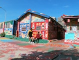 A vibrantly painted house covered in colorful artwork, including characters, symbols, and text in various languages. The building features a sloping roof decorated with multi-colored tiles, and the surrounding pavement also displays intricate designs. Two people are in front of the house, engaged in conversation, surrounded by the striking artistic details.