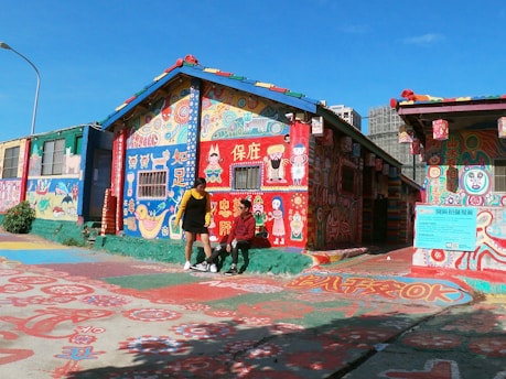 A vibrantly painted house covered in colorful artwork, including characters, symbols, and text in various languages. The building features a sloping roof decorated with multi-colored tiles, and the surrounding pavement also displays intricate designs. Two people are in front of the house, engaged in conversation, surrounded by the striking artistic details.
