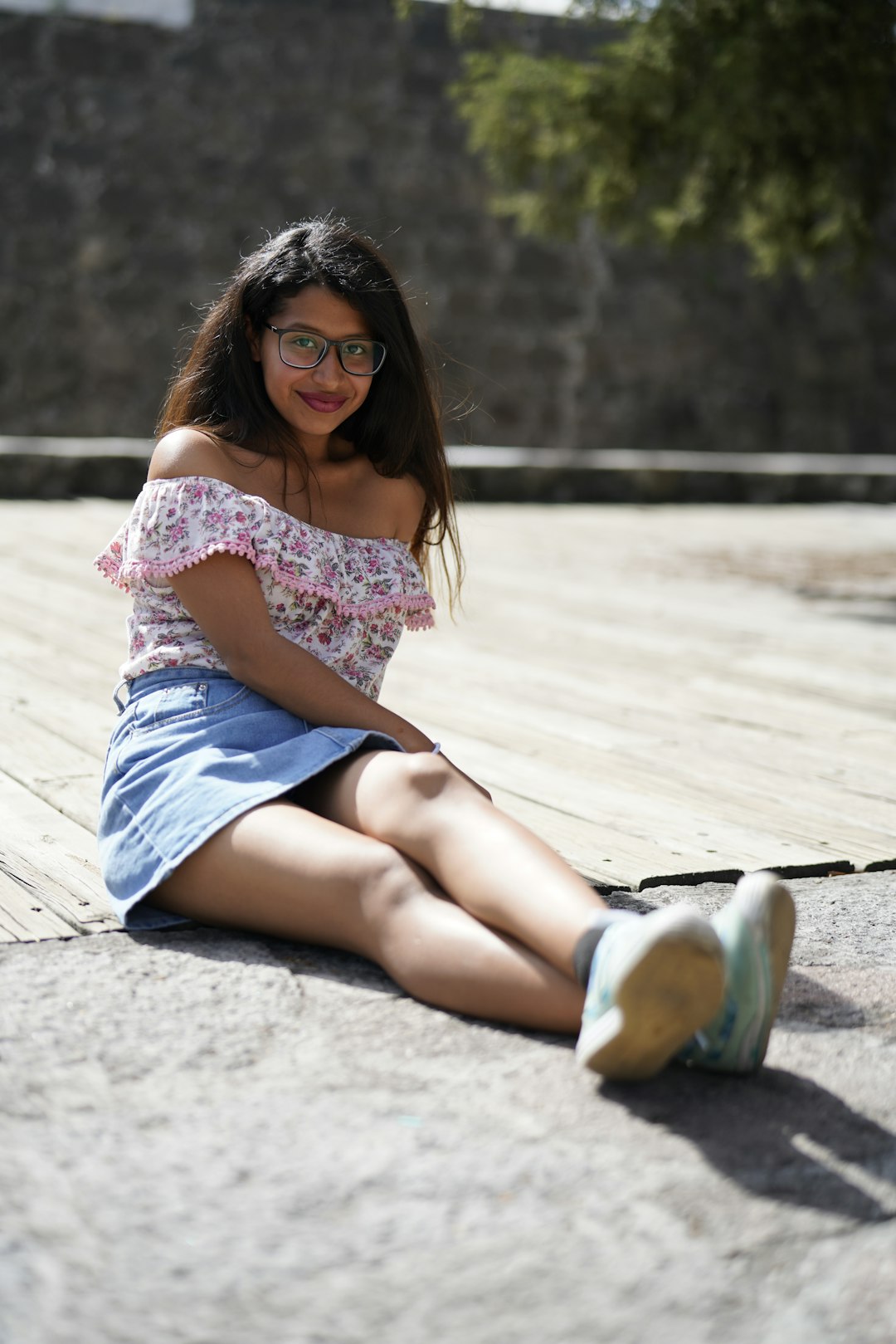 woman in white and pink floral shirt and blue denim shorts sitting on gray concrete floor