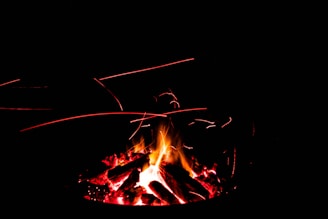 Logs burning with bright flames and red embers, surrounded by darkness. Sparks and glowing particles can be seen in the air, creating light trails.
