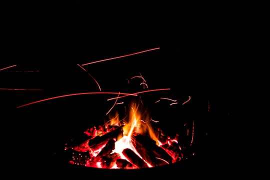Logs burning with bright flames and red embers, surrounded by darkness. Sparks and glowing particles can be seen in the air, creating light trails.