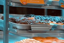 Assorted pastries, including apple fritters and chocolate-covered donuts, are displayed on metal racks in a bakery setting. The trays are organized neatly, showcasing the variety of sweet treats available.
