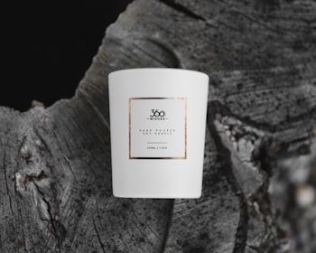 A white ceramic candle with a minimalist label, placed on the textured surface of a tree stump. The label reads '360 Wishes' and describes a hand-poured soy candle of 220ml capacity.