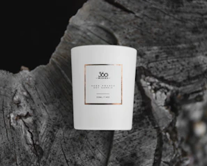 A white ceramic candle with a minimalist label, placed on the textured surface of a tree stump. The label reads '360 Wishes' and describes a hand-poured soy candle of 220ml capacity.