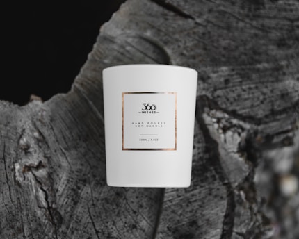 A white ceramic candle with a minimalist label, placed on the textured surface of a tree stump. The label reads '360 Wishes' and describes a hand-poured soy candle of 220ml capacity.