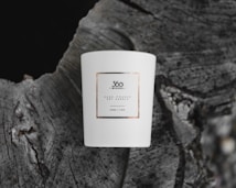 A white ceramic candle with a minimalist label, placed on the textured surface of a tree stump. The label reads '360 Wishes' and describes a hand-poured soy candle of 220ml capacity.