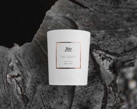 A white ceramic candle with a minimalist label, placed on the textured surface of a tree stump. The label reads '360 Wishes' and describes a hand-poured soy candle of 220ml capacity.
