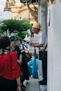 A group of people is gathered around an older man who seems to be giving a tour or explanation. The setting is outdoors near a temple-like structure with ornate architectural details and lush greenery in the background.