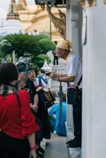 A group of people is gathered around an older man who seems to be giving a tour or explanation. The setting is outdoors near a temple-like structure with ornate architectural details and lush greenery in the background.