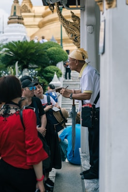 A group of people is gathered around an older man who seems to be giving a tour or explanation. The setting is outdoors near a temple-like structure with ornate architectural details and lush greenery in the background.