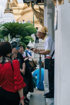 A group of people is gathered around an older man who seems to be giving a tour or explanation. The setting is outdoors near a temple-like structure with ornate architectural details and lush greenery in the background.