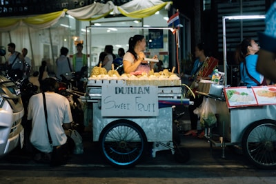 A street vendor is selling durians from a mobile cart. The cart has a sign displaying 'Durian Sweet Fruit' and is surrounded by other food carts. People are gathered around, some interacting with the vendor. The setting is busy and appears to be in an urban area at night, with artificial lighting from the carts.