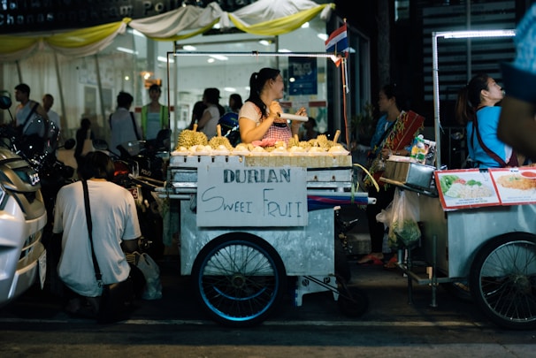 A street vendor is selling durians from a mobile cart. The cart has a sign displaying 'Durian Sweet Fruit' and is surrounded by other food carts. People are gathered around, some interacting with the vendor. The setting is busy and appears to be in an urban area at night, with artificial lighting from the carts.