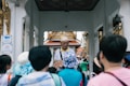 A group of tourists are gathered around a guide who is holding a brochure or map. The setting appears to be a historical or cultural site with ornate architecture in the background and an archway. The guide is wearing a hat and glasses, and the tourists are mostly wearing casual clothing and hats.