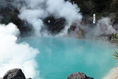 A vibrant turquoise hot spring with steam rising above the surface, surrounded by rocky terrain. Lush greenery can be seen around the edges, and a sign with Japanese characters is visible in the background.