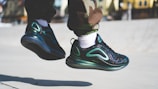 A pair of black and teal sneakers in mid-air against a bright background, worn by someone in camouflage pants and white socks. The sneakers have a sleek, reflective design.