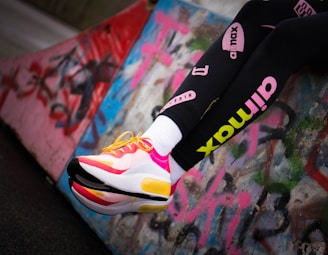 A person's legs wearing black leggings with colorful logos and bright, stylish sneakers. The sneakers are mainly white with pink and yellow accents and have a modern, sporty design. The background consists of a graffitied concrete surface, adding an urban and edgy feel.