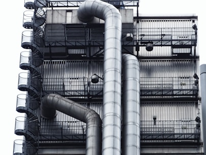 A close-up of an industrial structure featuring metal pipes and a series of metal stairs. The design is intricate with vertical and horizontal lines creating a grid-like pattern. The pipes curve smoothly, contrasting with the rigid, straight lines of the staircase railings.