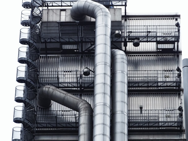 A close-up of an industrial structure featuring metal pipes and a series of metal stairs. The design is intricate with vertical and horizontal lines creating a grid-like pattern. The pipes curve smoothly, contrasting with the rigid, straight lines of the staircase railings.