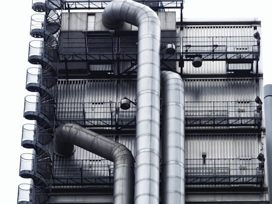 A close-up of an industrial structure featuring metal pipes and a series of metal stairs. The design is intricate with vertical and horizontal lines creating a grid-like pattern. The pipes curve smoothly, contrasting with the rigid, straight lines of the staircase railings.