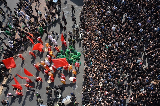 Aerial view of a crowded street with a large group of people dressed in black on the right side, contrasting with a colorful procession on the left featuring individuals in vibrant costumes with red and green tones and carrying red flags. The scene appears dynamic and energetic, suggesting a cultural or religious event.