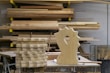 A woodworking shop with stacks of cut wooden boards organized on a surface. In the foreground, a uniquely shaped carved wooden piece stands upright. Behind, multiple planks of varying sizes are stored horizontally on racks.