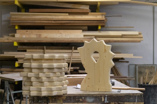 A woodworking shop with stacks of cut wooden boards organized on a surface. In the foreground, a uniquely shaped carved wooden piece stands upright. Behind, multiple planks of varying sizes are stored horizontally on racks.