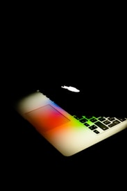A laptop partially open with a glowing apple logo on the back. The keyboard and touchpad display a spectrum of vibrant colors, creating a visually striking effect against the dark background.