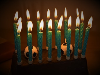 A cake is adorned with a series of lit blue candles that are arranged in an orderly manner. The flames flicker gracefully, casting a warm glow. Soccer ball decorations are nestled between the candles, emphasizing a celebratory theme related to soccer or sports.