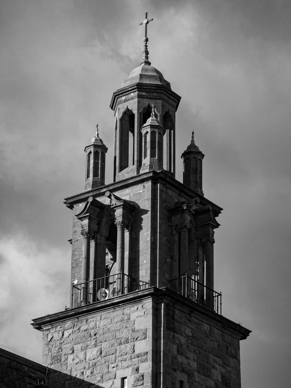 The image depicts a tall, stone church tower with four pointed spires and a cross at its peak. The tower is situated on a street corner, surrounded by an urban landscape. The photo is taken from a low angle, emphasizing the height and grandeur of the structure.