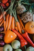 A colorful assortment of fresh vegetables and fruit, including carrots with green tops, root vegetables, an orange and red pepper, apples, onions, strawberries, and leafy greens.