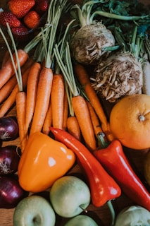 A colorful assortment of fresh vegetables and fruit, including carrots with green tops, root vegetables, an orange and red pepper, apples, onions, strawberries, and leafy greens.