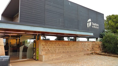 A modern building with a sleek, dark facade featuring the logo and name 'Telef&egrave;ric de Montju&iuml;c'. The building has large glass windows and is adjacent to a stone wall with greenery nearby. A cobblestone pathway leads up to the entrance.