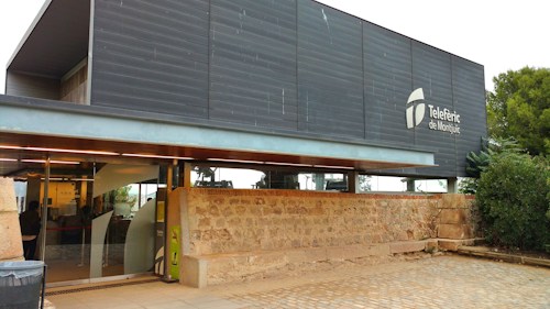 A modern building with a sleek, dark facade featuring the logo and name 'Telef&egrave;ric de Montju&iuml;c'. The building has large glass windows and is adjacent to a stone wall with greenery nearby. A cobblestone pathway leads up to the entrance.