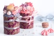 Two elegantly decorated cakes wrapped in floral-patterned paper, adorned with colorful meringue kisses and macarons on top. The cakes are tied with pastel ribbons, and there are scattered meringue kisses and small pearls on the white surface around them.